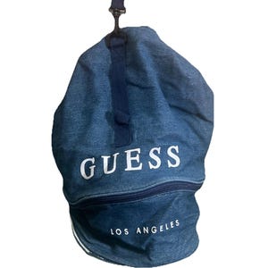 Può includere: Una borsa blu in stile denim con coulisse, con la scritta "GUESS" in grandi lettere bianche. Sotto il marchio, il testo "LOS ANGELES" è stampato in bianco. La borsa ha un cinturino e una cerniera blu scuro.