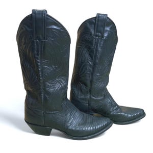 May include: A pair of black cowboy boots with a pointed toe and decorative stitching. The boots have a textured finish and a stacked heel. The shaft of the boots is tall, reaching up the calf. The boots are made of leather.