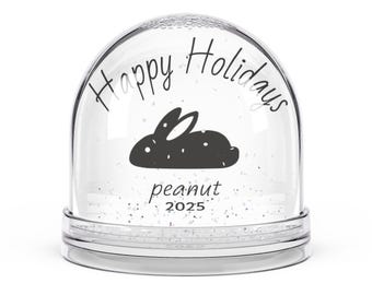 Op de afbeelding: Een heldere sneeuwbol met een zwart konijn silhouet en de woorden "Happy Holidays" en "peanut 2025" in zwart schrift. De bol is gevuld met glitter en staat op een heldere basis.