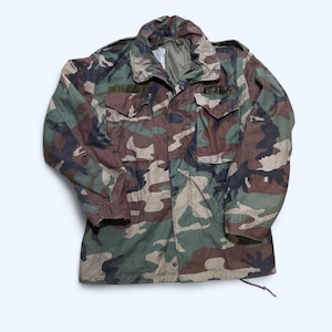 May include: Camouflage pattern jacket with a button-up front, two chest pockets, and two lower pockets. The jacket features a collar and the text "U.S. ARMY" on the right chest. The camouflage design includes shades of green, brown, and black.