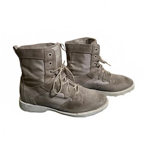 May include: A pair of gray combat boots with a suede and canvas design. The boots have a lace-up closure, a padded collar, and a durable rubber sole. The boots are designed for outdoor activities and rugged terrain.