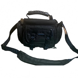 May include: A black camera bag featuring a top handle with a textured grip, a front zippered compartment, and two buckle closures with blue patterned webbing. The bag includes an adjustable shoulder strap with a padded section and blue accents.