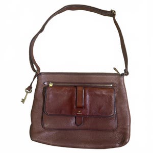 May include: Brown leather Fossil crossbody bag with a shoulder strap. The bag features a front pocket with a flap and a zipper closure. A gold-colored key charm hangs from the side. The bag has a textured surface.