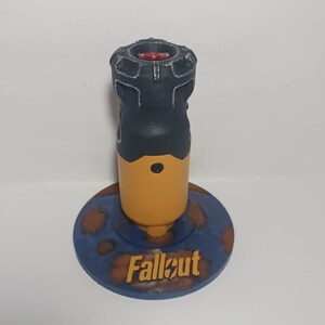 May include: A Fallout-themed bottle stopper. The stopper has a black top with a red centre, a yellow midsection, and a blue base with the word "Fallout" in gold lettering. The base has brown rust-like spots.