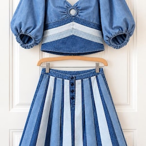May include: A two-piece denim outfit consisting of a cropped top and a skirt. The top features puff sleeves, a sweetheart neckline with a central ring detail, and chevron-patterned light and dark blue panels. The skirt has vertical light and dark blue stripes with a front button closure.