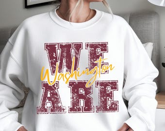 Op de afbeelding: Witte sweatshirt met de tekst "WE ARE Washington" in bordeauxrood en geel. De tekst heeft een distressed, vintage uitstraling. De sweatshirt is een crew neck stijl.