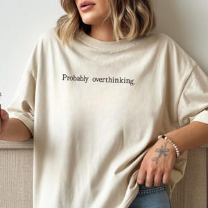 Puede incluir: Camiseta beige de gran tamaño con la frase "Probably overthinking" en negro. La camiseta se combina con vaqueros azules. Una persona sostiene una taza de café con una funda marrón.