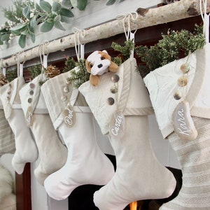 7 coordinating, but unique white Christmas stockings in cottage or farmhouse style are shown styled and hanging from a birch branch on a mantel. All the stockings have  agate name tags hanging from the buttons on their cuffs.