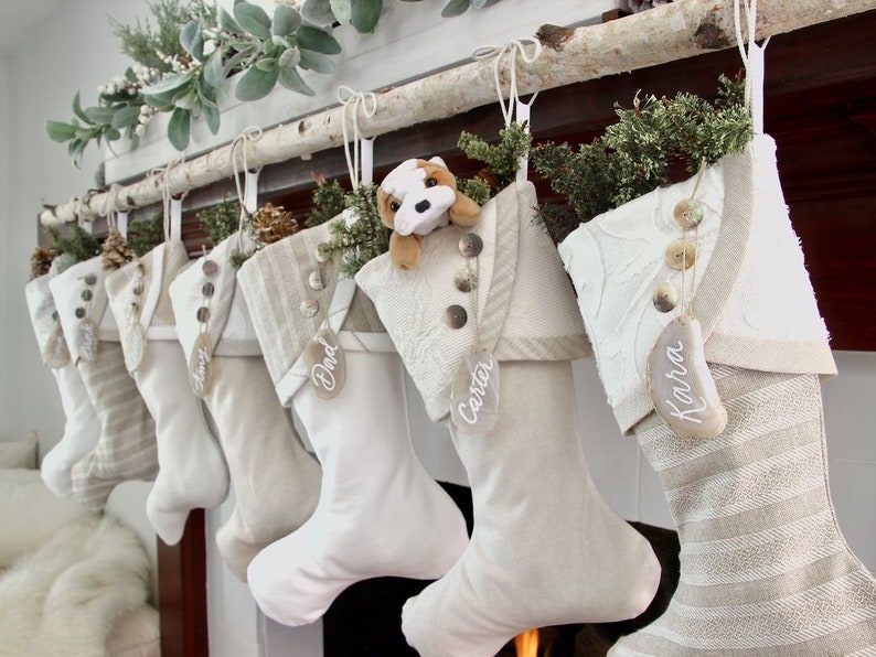 7 coordinating, but unique white Christmas stockings in cottage or farmhouse style are shown styled and hanging from a birch branch on a mantel. All the stockings have  agate name tags hanging from the buttons on their cuffs.