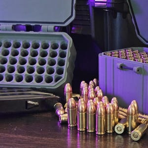 K&ouml;nnte beinhalten: Ein Bild von Messingpatronen und Munitionsaufbewahrungsboxen. Ein gr&uuml;ner Beh&auml;lter ist mit Patronen gef&uuml;llt, weitere sind davor verstreut. Die Beh&auml;lter sind ge&ouml;ffnet und geben den Inhalt frei. Die Patronen haben eine gl&auml;nzende goldene Farbe.