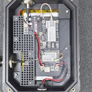 May include: A black rectangular electronic device with an open top, revealing internal components. The device includes circuit boards, wiring, and a black antenna connector. The text "RAK13302" is visible on one of the boards.
