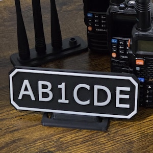 May include: A black and white sign with the text "AB1CDE" is displayed in front of several black handheld radios and antennas. The radios have buttons and screens. The items are on a brown wooden surface.