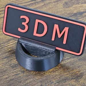 May include: A black and red 3D printed sign with the letters "3DM" in orange. The sign is rectangular and sits in a black curved holder. The background is a wooden surface.