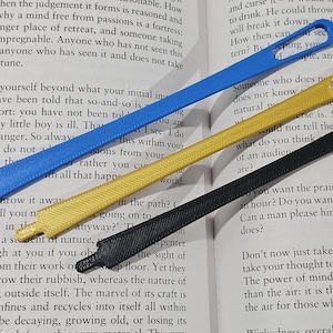 May include: Three ribbon threaders in blue, gold, and black lie on an open book. Each threader has a loop at one end and a flat, textured tip at the other. The book's pages display printed text.