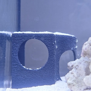 May include: Two dark blue plastic aquarium decorations with multiple holes. The decorations are sitting on a white sand substrate.