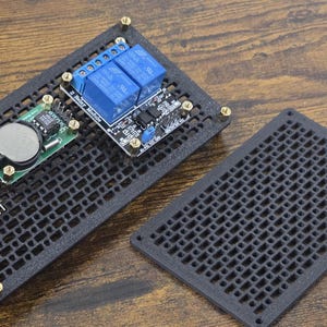 May include: A black circuit board with various electronic components, including blue relays, a round battery, and a blue sensor. The board is mounted on a black, perforated base with a matching cover. The components are secured with gold-colored screws.