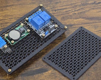 Könnte beinhalten: Eine schwarze Leiterplatte mit verschiedenen elektronischen Bauteilen, darunter blaue Relais, eine runde Batterie und ein blauer Sensor. Die Platine ist auf einer schwarzen, perforierten Basis mit passendem Deckel montiert. Die Komponenten sind mit goldfarbenen Schrauben befestigt.