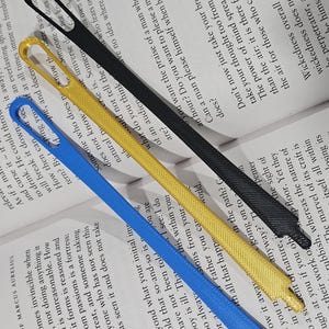 May include: Four colorful ribbon bookmarks: blue, yellow, black, and a partially visible white one. The bookmarks are lying on top of an open book with text. The bookmarks are made of a woven material.