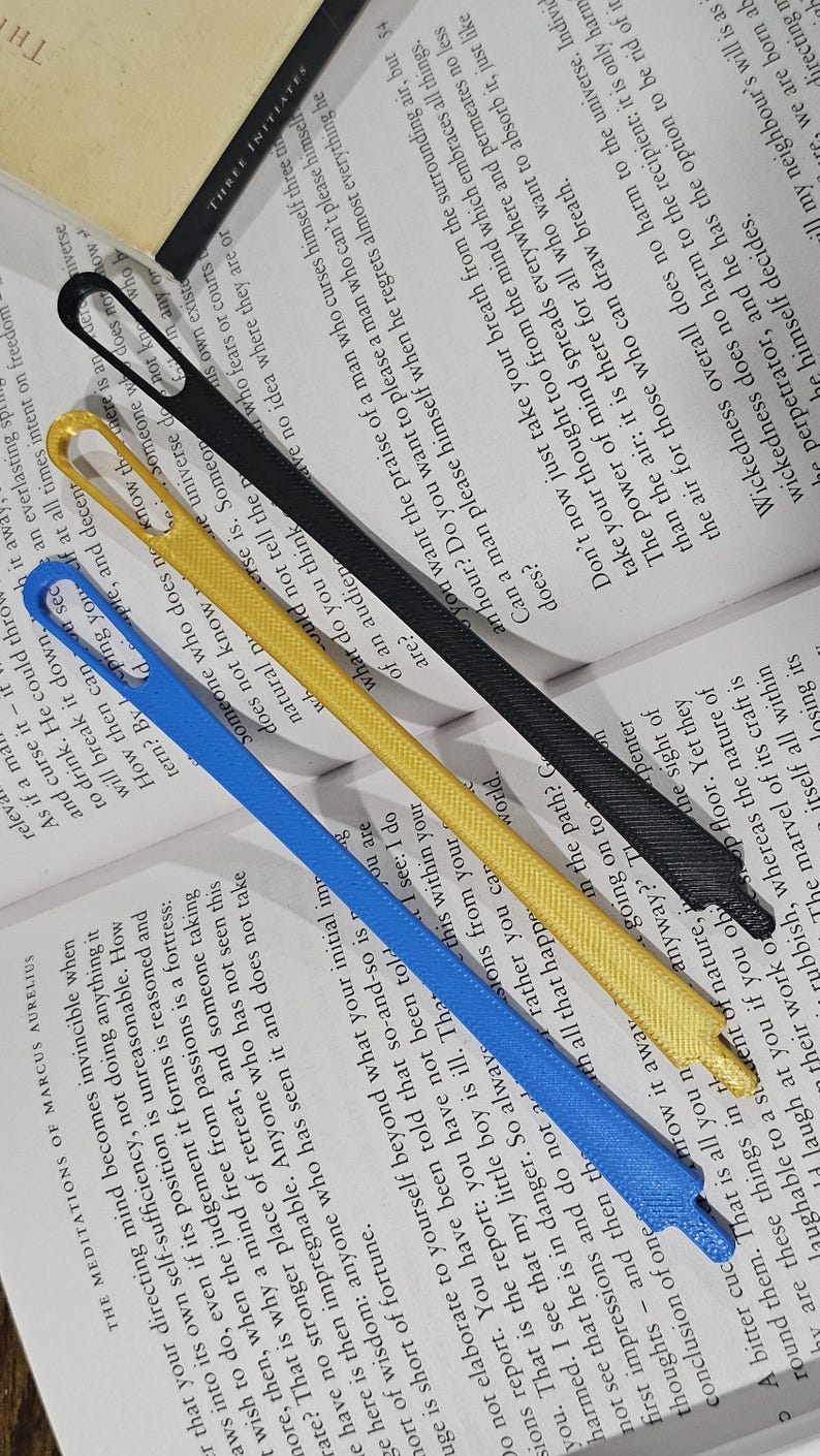 May include: Four colorful ribbon bookmarks: blue, yellow, black, and a partially visible white one. The bookmarks are lying on top of an open book with text. The bookmarks are made of a woven material.
