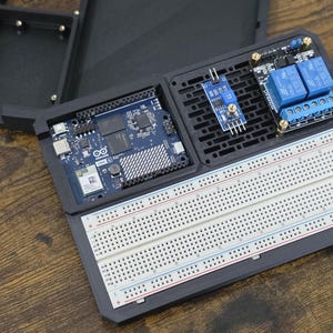 May include: A black electronic project enclosure with an Arduino board, breadboard, and various electronic components. The components include blue relays and a blue sensor module. The enclosure is open, revealing the internal components.