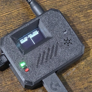 May include: A black electronic device with a small screen displaying data. The device has a textured surface, a green and red button, and a USB connector. A black cable is connected to the device.