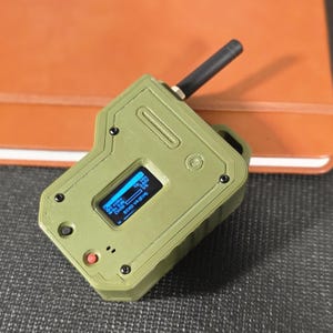 May include: An olive green electronic device with a small screen displaying text and graphics. It has a black antenna, a red button, and several black screws. The device appears to be a handheld gadget, possibly for data collection or control.