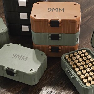 K&ouml;nnte beinhalten: Eine Sammlung von Munitionsaufbewahrungsboxen in verschiedenen Farben, darunter Gr&uuml;n, Braun und Schwarz. Die Boxen sind mit "9MM" und "5.56MM ECHO" beschriftet. Eine Box ist ge&ouml;ffnet und zeigt zahlreiche messingfarbene Kugeln.