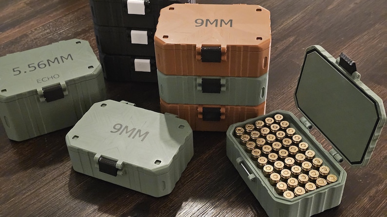 K&ouml;nnte beinhalten: Eine Sammlung von Munitionsaufbewahrungsboxen in verschiedenen Farben, darunter Gr&uuml;n, Braun und Schwarz. Die Boxen sind mit "9MM" und "5.56MM ECHO" beschriftet. Eine Box ist ge&ouml;ffnet und zeigt zahlreiche messingfarbene Kugeln.