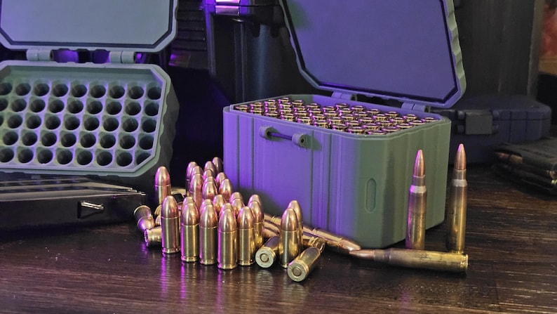 K&ouml;nnte beinhalten: Ein Bild von Messingpatronen und Munitionsaufbewahrungsboxen. Ein gr&uuml;ner Beh&auml;lter ist mit Patronen gef&uuml;llt, weitere sind davor verstreut. Die Beh&auml;lter sind ge&ouml;ffnet und geben den Inhalt frei. Die Patronen haben eine gl&auml;nzende goldene Farbe.