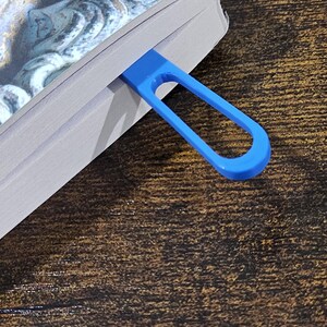 May include: A blue, loop-shaped bookmark is inserted into a book. The bookmark is made of a solid, smooth material. The book has a dark cover with a detailed image. The background is a dark wood grain.