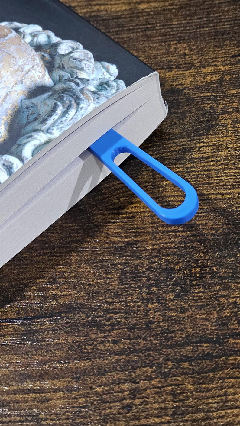 May include: A blue, loop-shaped bookmark is inserted into a book. The bookmark is made of a solid, smooth material. The book has a dark cover with a detailed image. The background is a dark wood grain.