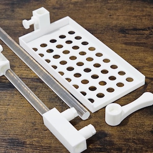 May include: A collection of white 3D-printed components, including a slotted tray, clear tubes, and various mounting brackets. The items are arranged on a dark wood-grain surface, suggesting a scientific or technical theme.