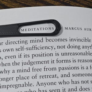May include: A black bookmark with the word "MEDITATIONS" printed on it, resting on a book page. The book is open, and the text "MARCUS AURELIUS" is visible. The bookmark is made of a textured material.