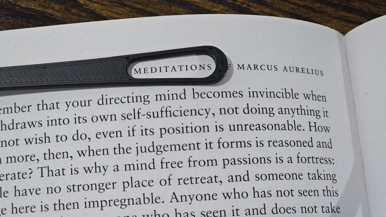 May include: A black bookmark with the word "MEDITATIONS" printed on it, resting on a book page. The book is open, and the text "MARCUS AURELIUS" is visible. The bookmark is made of a textured material.