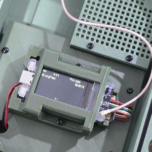 May include: An open, olive-green electronic device enclosure with a small screen displaying text. The device has a grid-like component and various wires and connectors. The text on the screen reads "LongFast" and "No SMS".