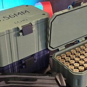 K&ouml;nnte beinhalten: Zwei olivgr&uuml;ne Munitionsbeh&auml;lter, einer davon ge&ouml;ffnet, gef&uuml;llt mit Patronen mit Messingh&uuml;lsen. Der obere Beh&auml;lter ist mit "5.56MM ECHO" beschriftet. Die Beh&auml;lter sind auf einem dunkelblauen Beh&auml;lter gestapelt.