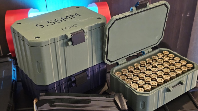 K&ouml;nnte beinhalten: Zwei olivgr&uuml;ne Munitionsbeh&auml;lter, einer davon ge&ouml;ffnet, gef&uuml;llt mit Patronen mit Messingh&uuml;lsen. Der obere Beh&auml;lter ist mit "5.56MM ECHO" beschriftet. Die Beh&auml;lter sind auf einem dunkelblauen Beh&auml;lter gestapelt.