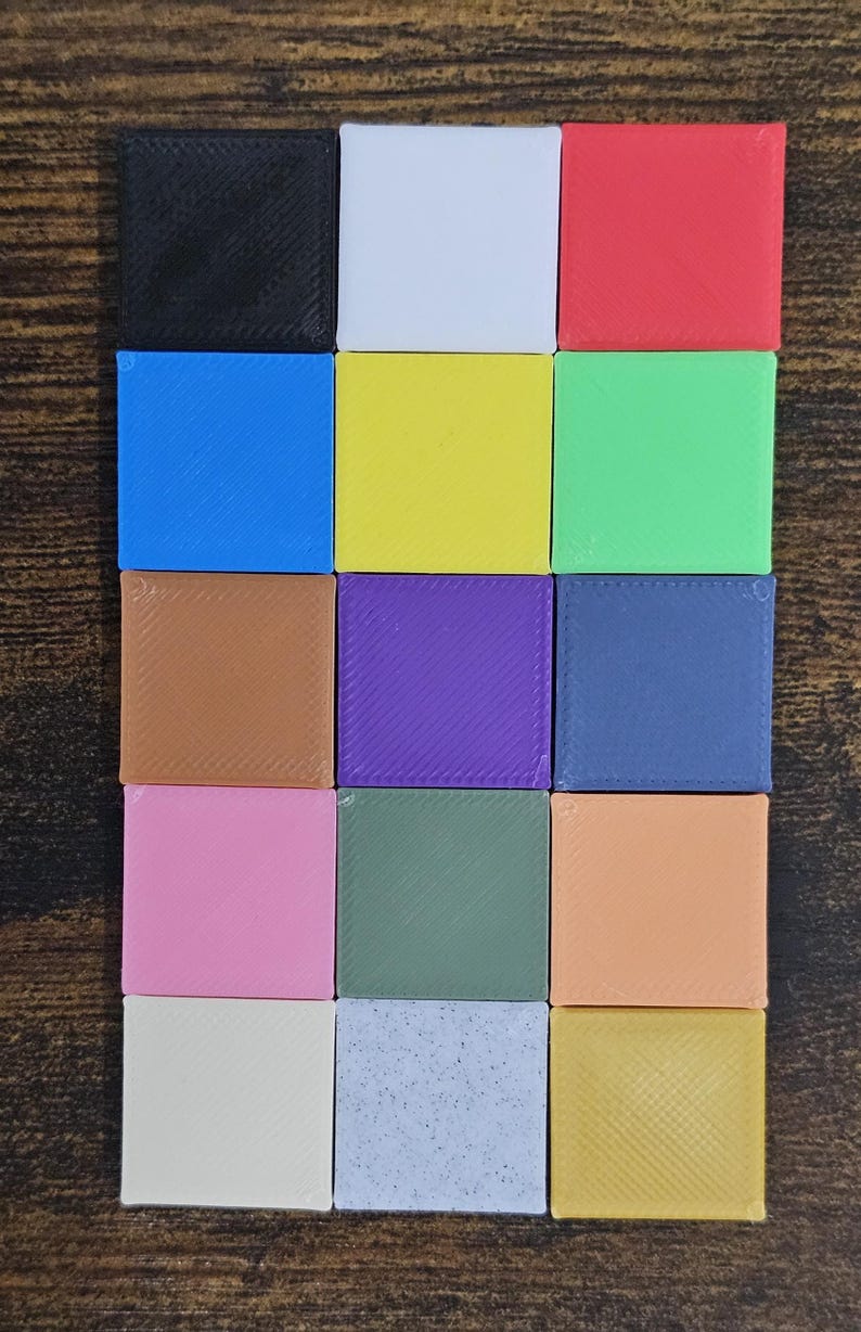 May include: A collection of sixteen small, square blocks in various colors, including black, white, red, blue, yellow, green, brown, purple, and pink. The blocks are arranged in a grid pattern on a wooden surface.
