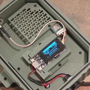 May include: An open, olive-green electronic enclosure with internal components. A small screen displays text. Wires and connectors are visible, including a coaxial cable and a small circuit board. The enclosure has a grid-like vent.