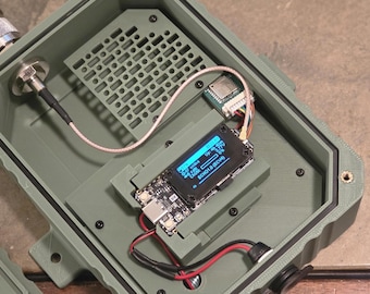May include: An open, olive-green electronic enclosure with internal components. A small screen displays text. Wires and connectors are visible, including a coaxial cable and a small circuit board. The enclosure has a grid-like vent.