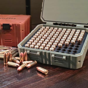 Puede incluir: Una caja de munición verde claro abierta llena de balas con casquillos de latón. Detrás hay una segunda caja naranja cerrada con el texto "5.56MM". Las balas sueltas están esparcidas sobre una superficie de madera.