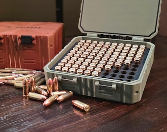 May include: An open, light green ammunition case filled with brass-cased bullets. A second, closed orange case with the text "5.56MM" is behind it. Loose bullets are scattered on a wooden surface.