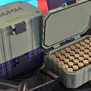 K&ouml;nnte beinhalten: Zwei olivgr&uuml;ne Munitionsbeh&auml;lter, einer davon ge&ouml;ffnet und mit messingfarbenen Kugeln gef&uuml;llt. Der geschlossene Beh&auml;lter tr&auml;gt die Aufschrift "5.56MM ECHO" auf dem Deckel. Die Beh&auml;lter sind aus einem robusten Material gefertigt.