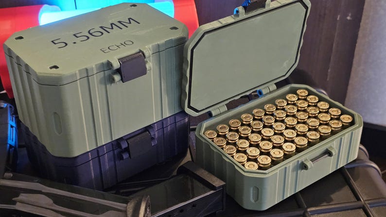 K&ouml;nnte beinhalten: Zwei olivgr&uuml;ne Munitionsbeh&auml;lter, einer davon ge&ouml;ffnet und mit messingfarbenen Kugeln gef&uuml;llt. Der geschlossene Beh&auml;lter tr&auml;gt die Aufschrift "5.56MM ECHO" auf dem Deckel. Die Beh&auml;lter sind aus einem robusten Material gefertigt.