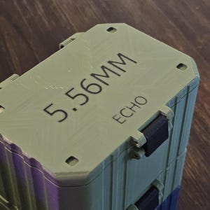 K&ouml;nnte beinhalten: Ein gr&uuml;ner und violetter rechteckiger Beh&auml;lter mit dem Text "5.56MM ECHO" auf der Oberseite. Der Beh&auml;lter hat schwarze Verschl&uuml;sse und steht auf einer Holzoberfl&auml;che.