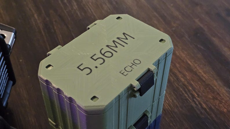K&ouml;nnte beinhalten: Ein gr&uuml;ner und violetter rechteckiger Beh&auml;lter mit dem Text "5.56MM ECHO" auf der Oberseite. Der Beh&auml;lter hat schwarze Verschl&uuml;sse und steht auf einer Holzoberfl&auml;che.