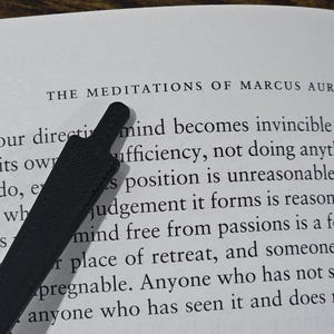 May include: Open book with the title "THE MEDITATIONS OF MARCUS AURELIUS" visible. A black bookmark is placed diagonally across the page. The text is in black font on a white background.