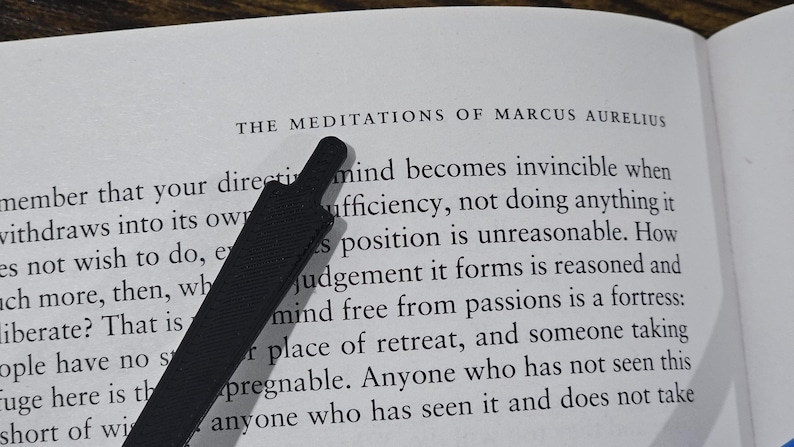 May include: Open book with the title "THE MEDITATIONS OF MARCUS AURELIUS" visible. A black bookmark is placed diagonally across the page. The text is in black font on a white background.
