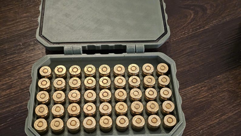 K&ouml;nnte beinhalten: Ein gr&uuml;ner, rechteckiger Koffer mit einem Klappdeckel, gef&uuml;llt mit Reihen von messingfarbener Munition. Der Koffer ist ge&ouml;ffnet und zeigt die ordentlich angeordneten Kugeln. Der Koffer besteht aus einem strukturierten Material.