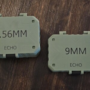 K&ouml;nnte beinhalten: Zwei olivgr&uuml;ne, rechteckige Beh&auml;lter mit Klappdeckeln. Der gr&ouml;&szlig;ere Beh&auml;lter ist mit "5.56MM" und "ECHO" beschriftet. Der kleinere Beh&auml;lter ist mit "9MM" und "ECHO" beschriftet. Beide liegen auf einer Holzoberfl&auml;che.
